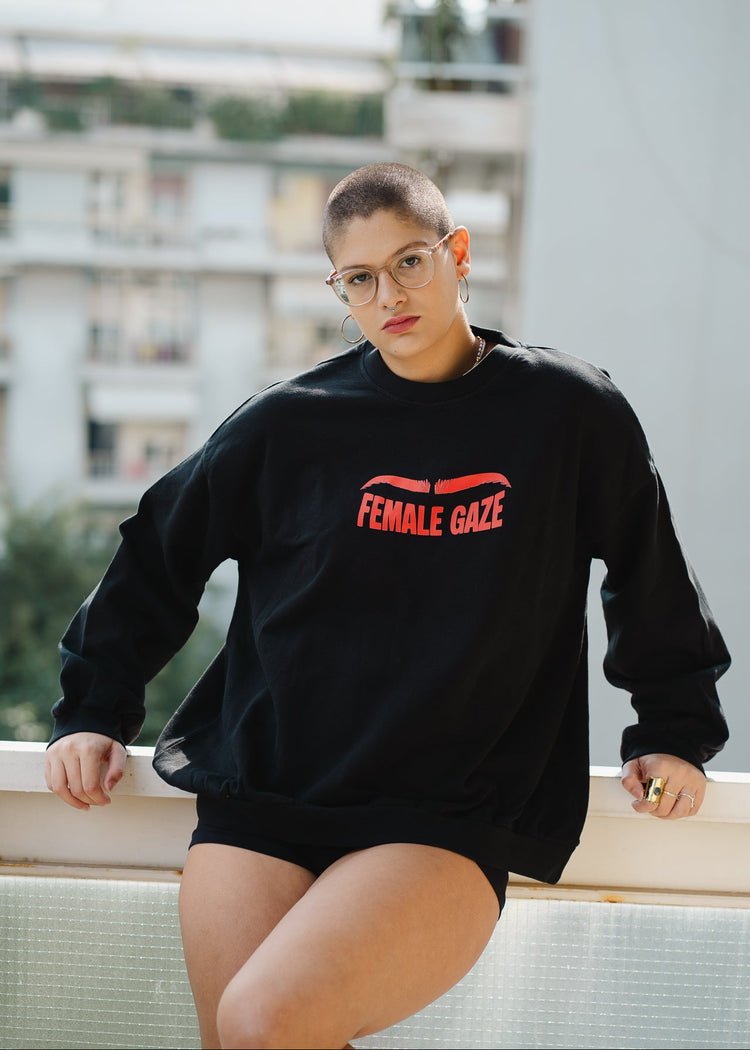 Person wearing a black sweatshirt with 'FEMALE GAZE' text, sitting on a ledge.