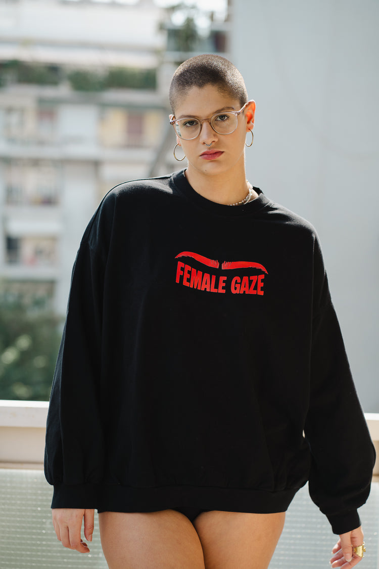 Person wearing a black sweatshirt with 'FEMALE GAZE' text, standing outdoors.