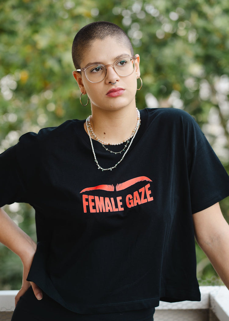 Person wearing a black t-shirt with 'FEMALE GAZE' text, standing outdoors.