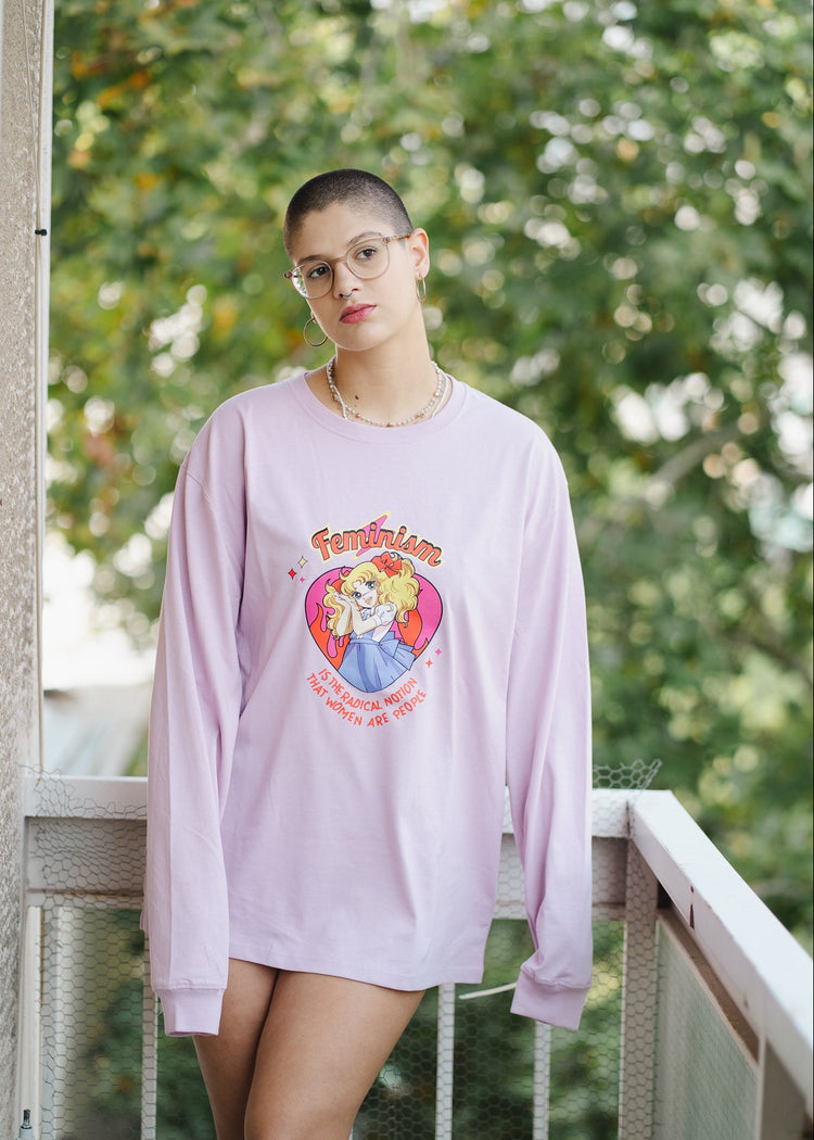 Person wearing a light purple oversized sweatshirt with a graphic design, standing on a balcony with greenery in the background.