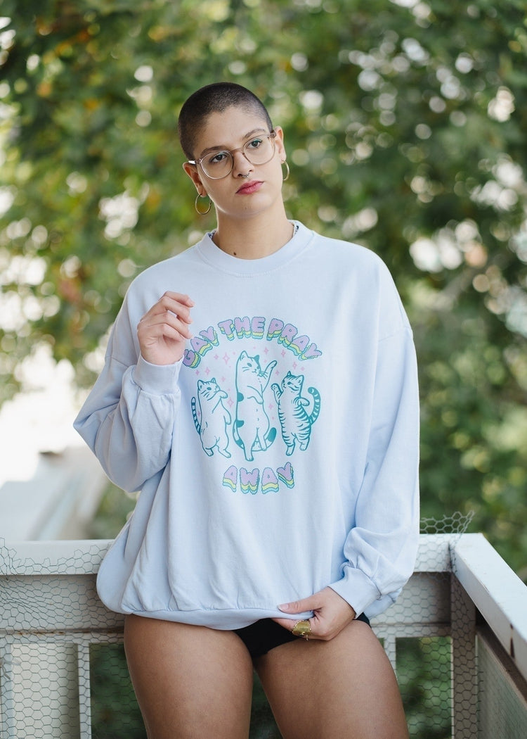 Person wearing a light blue sweatshirt with graphic design, standing outdoors with greenery in the background