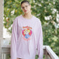 Person wearing a light purple oversized sweatshirt with a graphic design, standing on a balcony with greenery in the background.