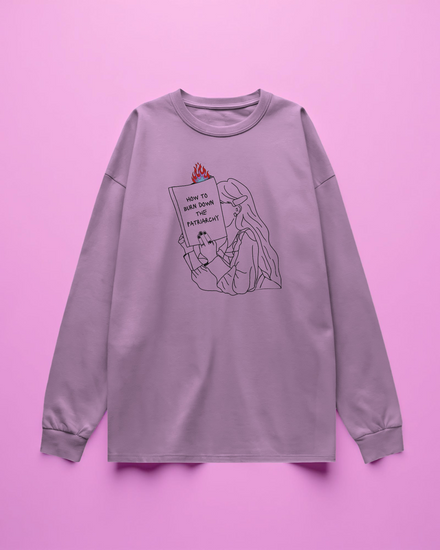 pink long sleeve t-shirt with graphic design of a person holding a book titled 'How to Burn Down the Patriarchy' on a purple background.