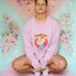 Person wearing a pink sweatshirt with a graphic design, sitting in front of a colorful floral wall.