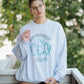 Person wearing a light blue sweatshirt with graphic design, standing outdoors with greenery in the background