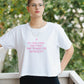 Person wearing a white t-shirt with pink text, standing outdoors with greenery in the background