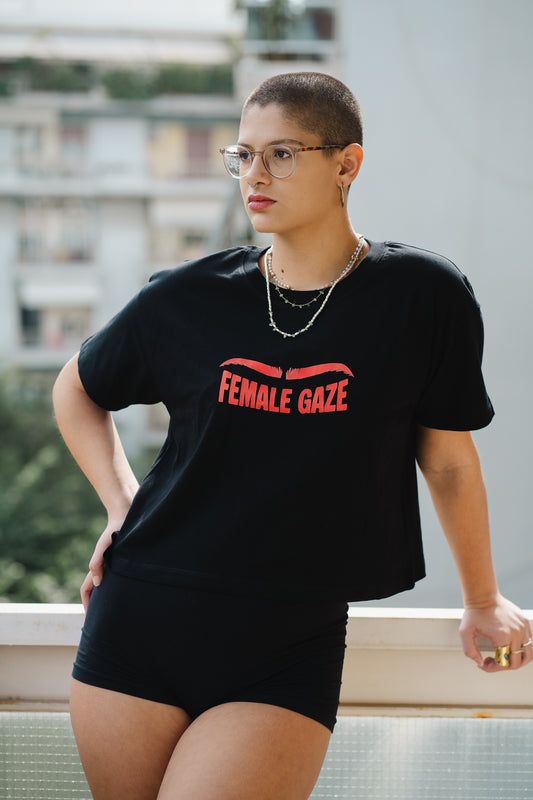 Person wearing a black t-shirt with 'FEMALE GAZE' text, standing outdoors.